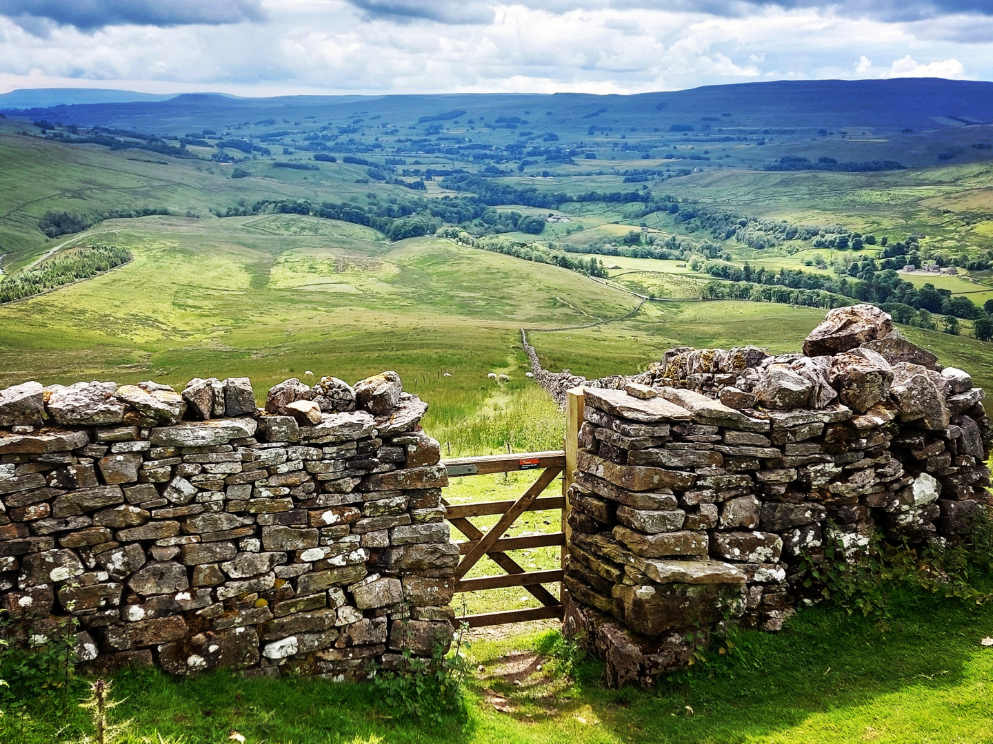 Views through a gate in a stone wall over the rolling green hills of Yorkshire, with its famous peaks in the far distance.